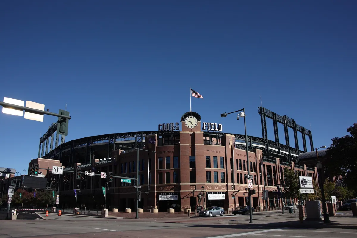 A wide exterior view of Coors Field stadium, complete with the American flag and a prominent clock tower. The area is animated with the excitement of crowds gathering for events, reflecting Denver's community spirit and event-hosting prowess.