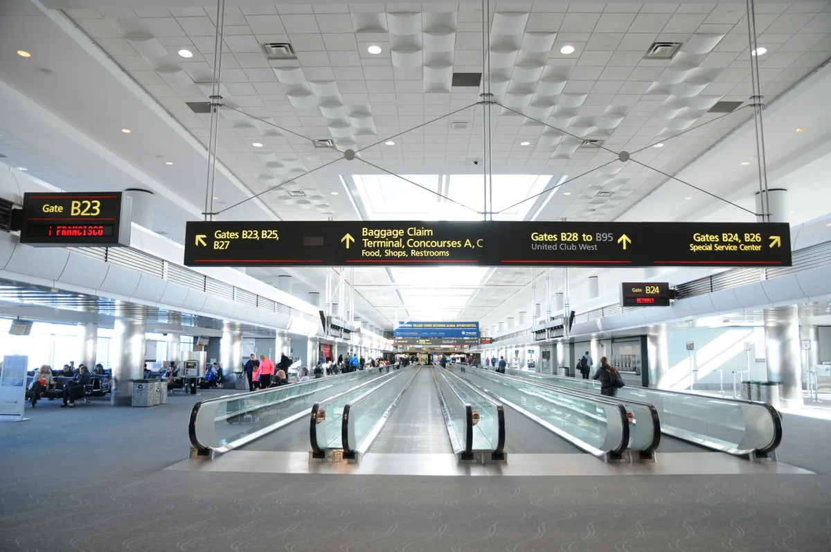 This image captures a bustling interior scene at an airport, featuring a moving walkway, passengers waiting with luggage, gate signs, and shops in the background. People appear energetic and excited, possibly embarking on or returning from group adventures, reunions, or fun getaways.