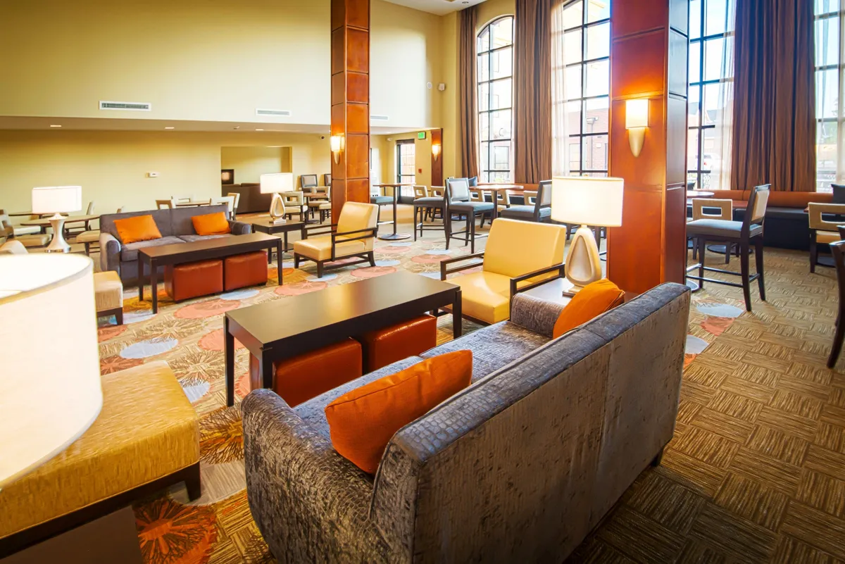 An open, sunlit living area with large windows, plush seating, and decorative lighting, creating a relaxed and welcoming setting. The space is designed for socializing, unwinding with a book, or staying productive on a laptop, symbolizing comfort and versatility.