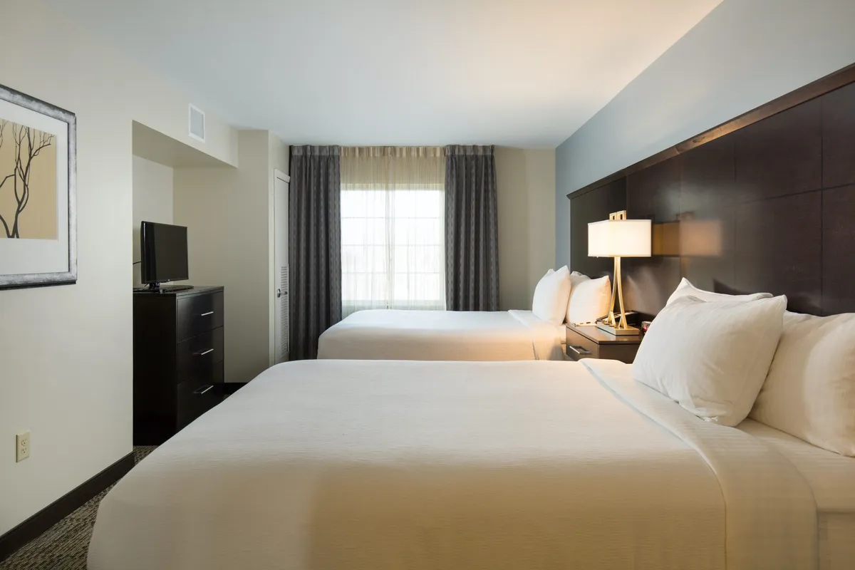 This image captures a bright, inviting bedroom with two beds, a window letting in natural light, and a nightstand with a lamp. The layout emphasizes space for multiple guests, a cozy atmosphere, and restful comfort.
