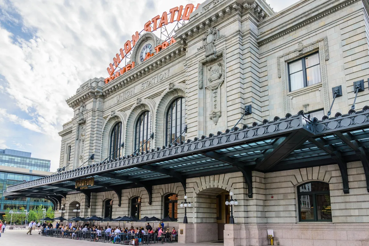 A dynamic exterior scene featuring a historic building with iconic neon signage, large windows, and an active outdoor seating area. The space is bustling with travelers, diners, and visitors, creating a lively, urban atmosphere.