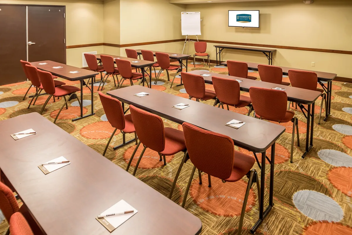 A well-lit event space with rows of tables, bold red chairs, and a prominent presentation screen, inviting guests to gather for meetings, workshops, or receptions. The layout encourages interaction and group activity.