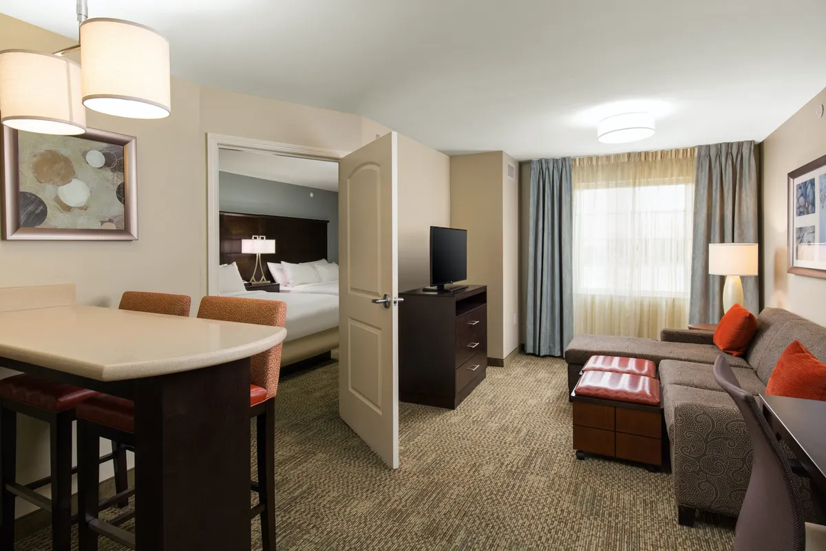 The image showcases a stylish, open-plan suite interior. A modern bar counter with stools invites morning coffee or casual meals, while a comfortable sofa provides space for relaxing or gathering with loved ones. The bedroom doorway hints at spacious accommodations, and a large window with soft curtains bathes the room in natural light, creating a warm, inviting atmosphere. A television offers entertainment options, and the overall scene feels perfect for unwinding after a day in Denver.