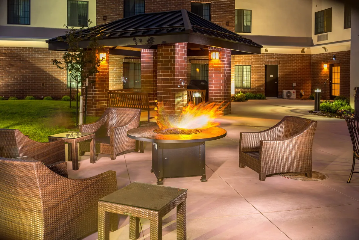 This exterior scene presents a welcoming fire pit surrounded by seating, set against the backdrop of the Staybridge Suites building. Guests are seen socializing and relaxing in a landscaped area, suggesting a cozy, communal atmosphere perfect for unwinding after a day of exploration.