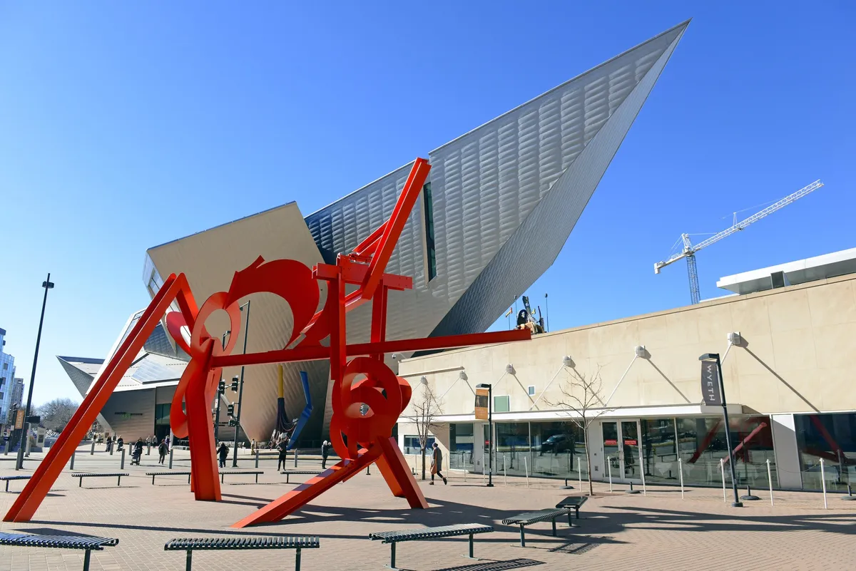 Set in a dynamic urban plaza, this image features a striking red sculpture and a modern architectural structure, with people leisurely strolling and engaging with public art. The setting buzzes with activity and culture, evoking the spirit of Denver’s vibrant attractions.