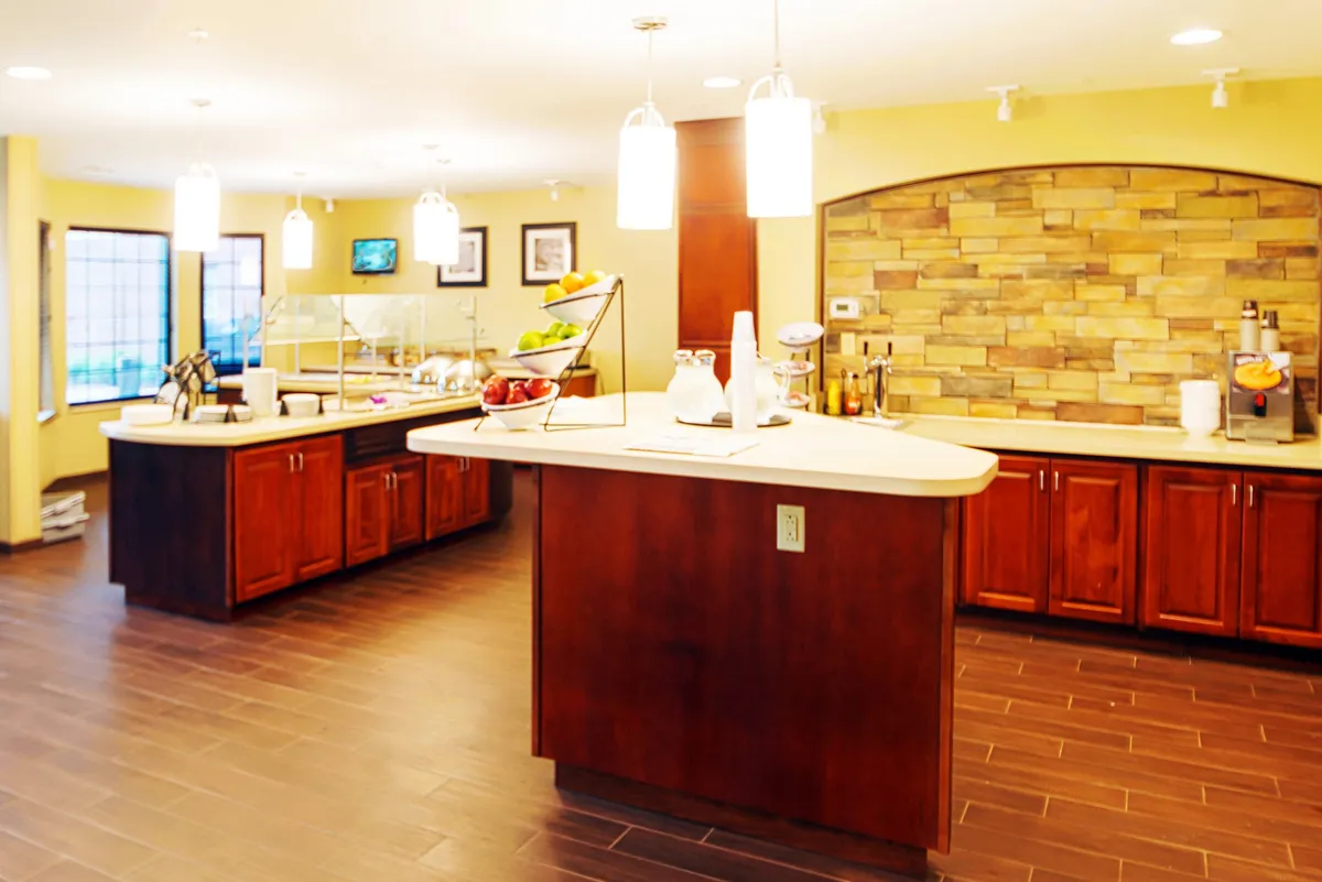 A stylish kitchen and dining area with a central island displaying fresh fruit, stone accents, warm wood cabinetry, and hanging light fixtures. The image evokes morning conversations, shared breakfasts, or casual socializing in a bright, friendly environment.