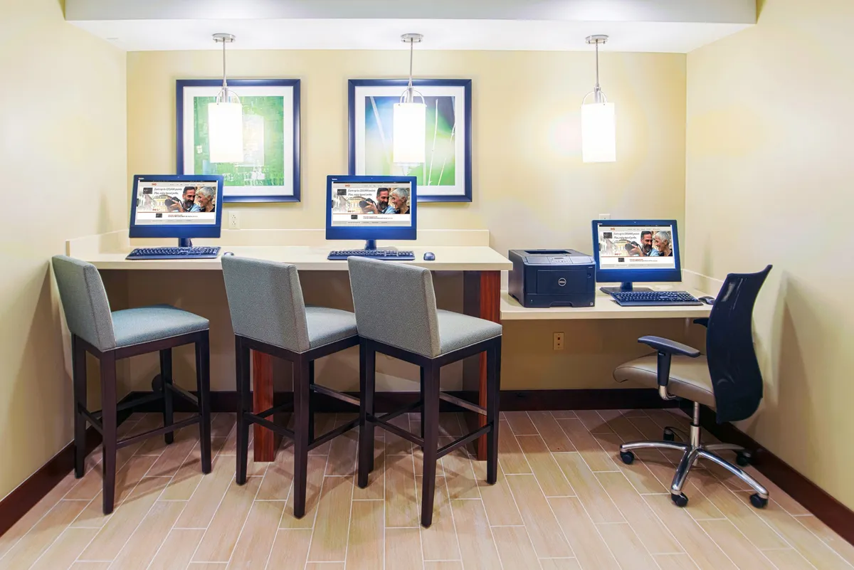 A modern business center with multiple computers on a sleek counter, bar stools for seating, a printer for document needs, and framed artwork on the walls. The space is well-lit and designed for productivity and collaboration.