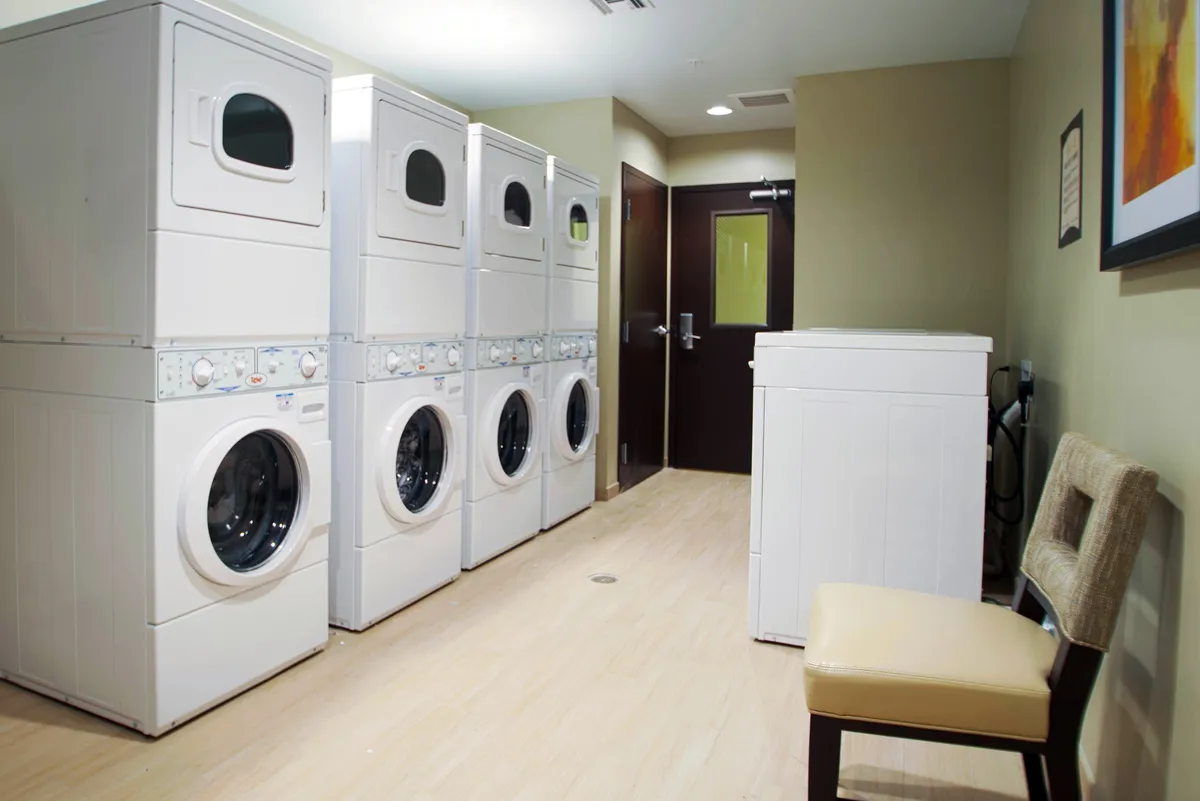 A practical on-site laundry facility featuring stacked washer and dryer units, a single washing machine, and a chair for waiting. The room is clean and efficiently organized, supporting extended-stay routines.