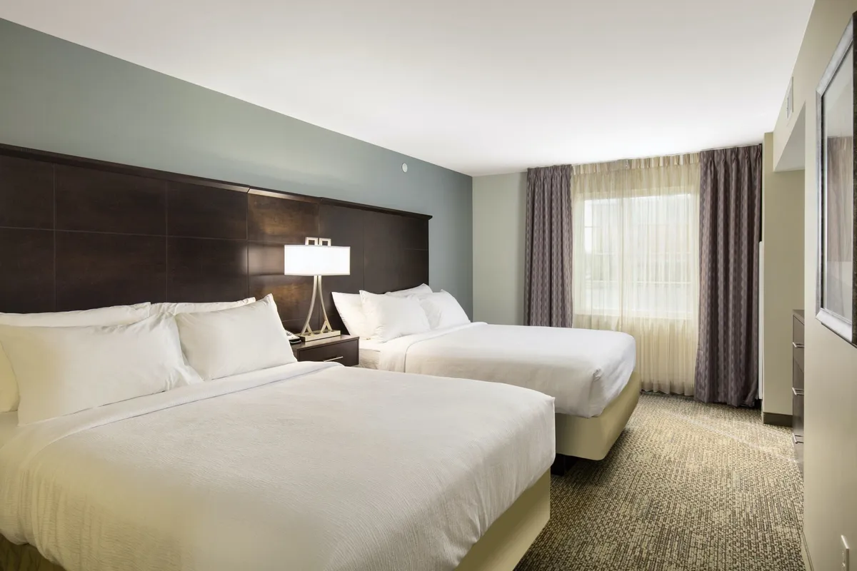 The bedroom scene features two beds, a nightstand with a lamp, and a window with curtains, conveying a peaceful and comfortable environment ideal for relaxation after a busy day.